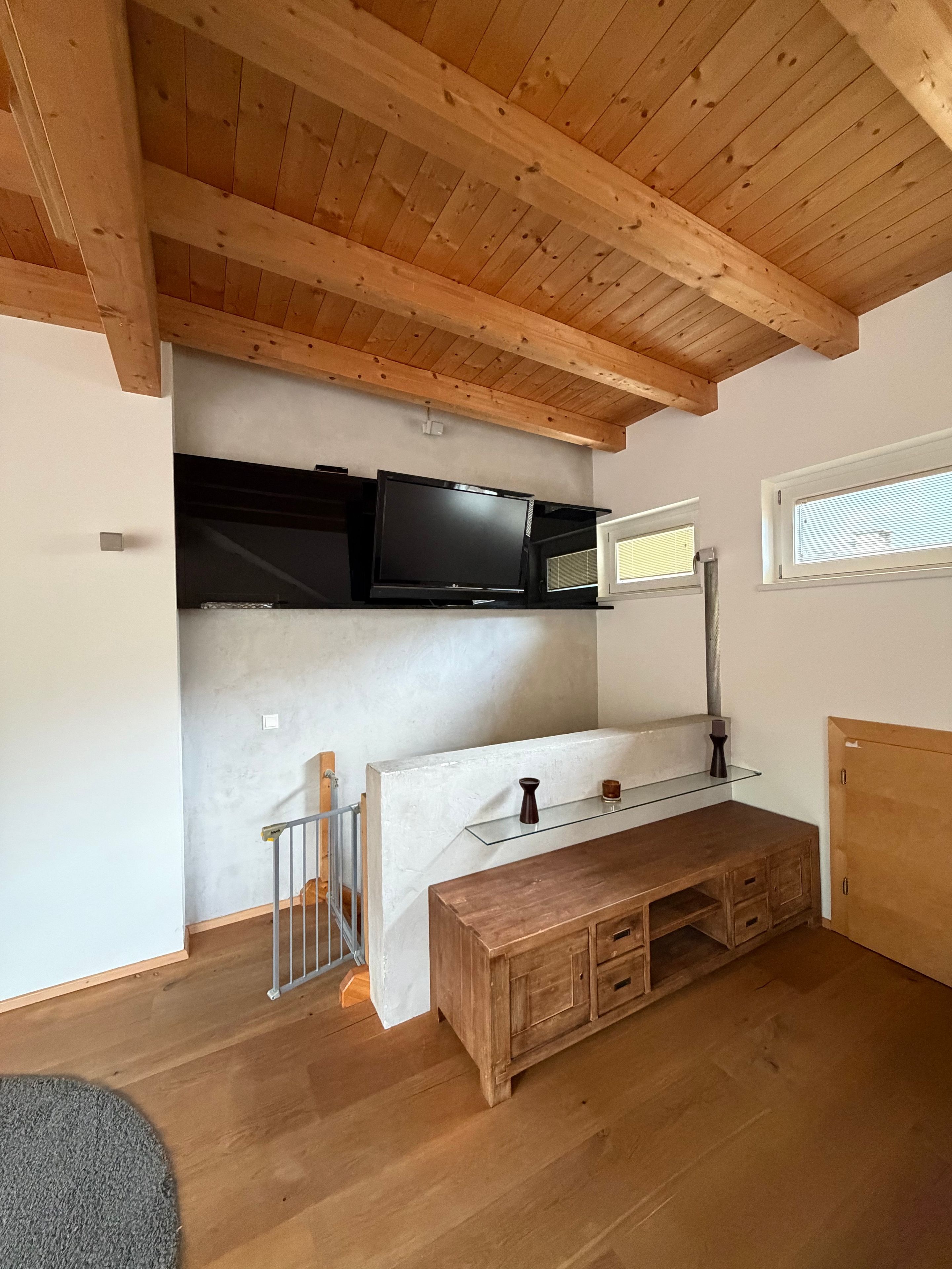 A room with a wooden ceiling, a TV mounted on a wall, a wooden bench, and a window with blinds.