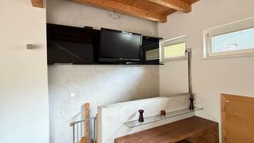A room with a wooden ceiling, a TV mounted on a wall, a wooden bench, and a window with blinds.