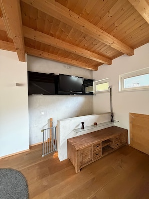 A room with a wooden ceiling, a TV mounted on a wall, a wooden bench, and a window with blinds.