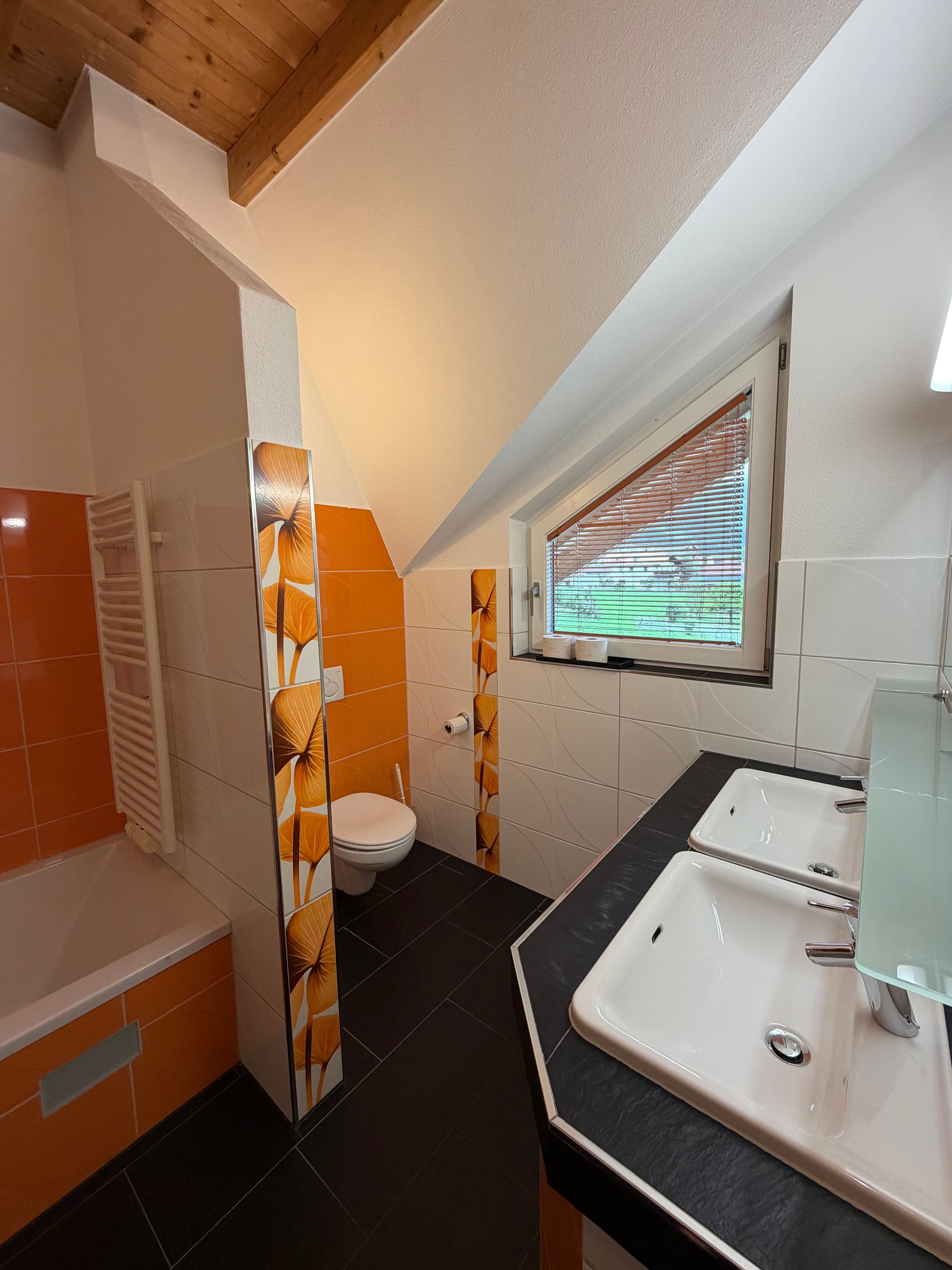 Ein Badezimmer mit Toilette, Waschbecken und Duschbereich, mit orangefarbenen und weißen Fliesen sowie einem Fenster mit Jalousien.