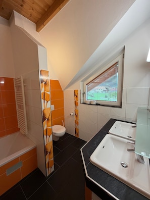 Een badkamer met toilet, wastafel en douchegedeelte, met oranje en witte tegels en een raam met zonwering.