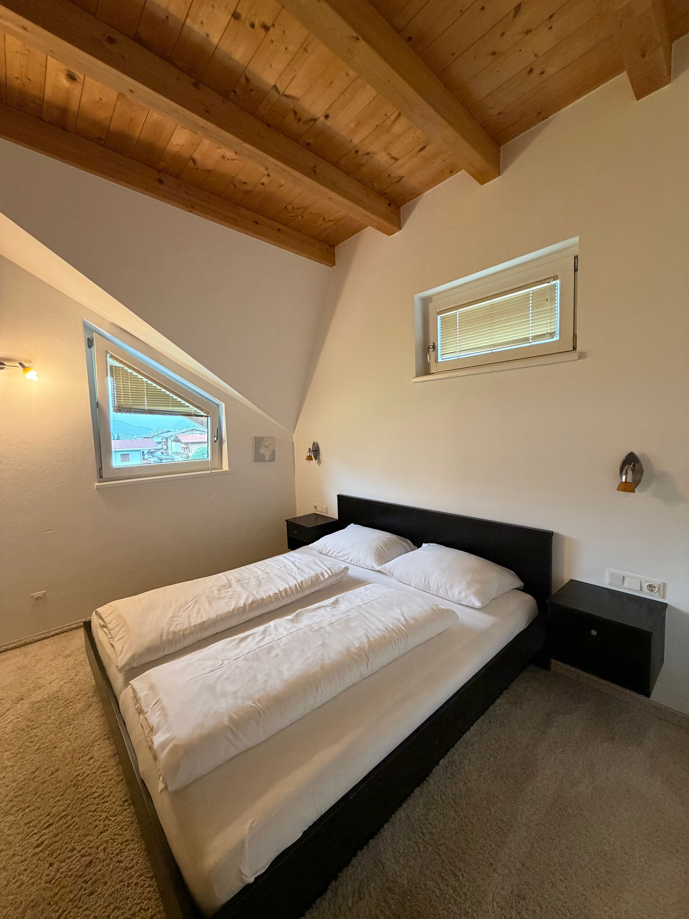 A bedroom with a wooden ceiling, a bed with white bedding, two bedside tables, and two windows with blinds.