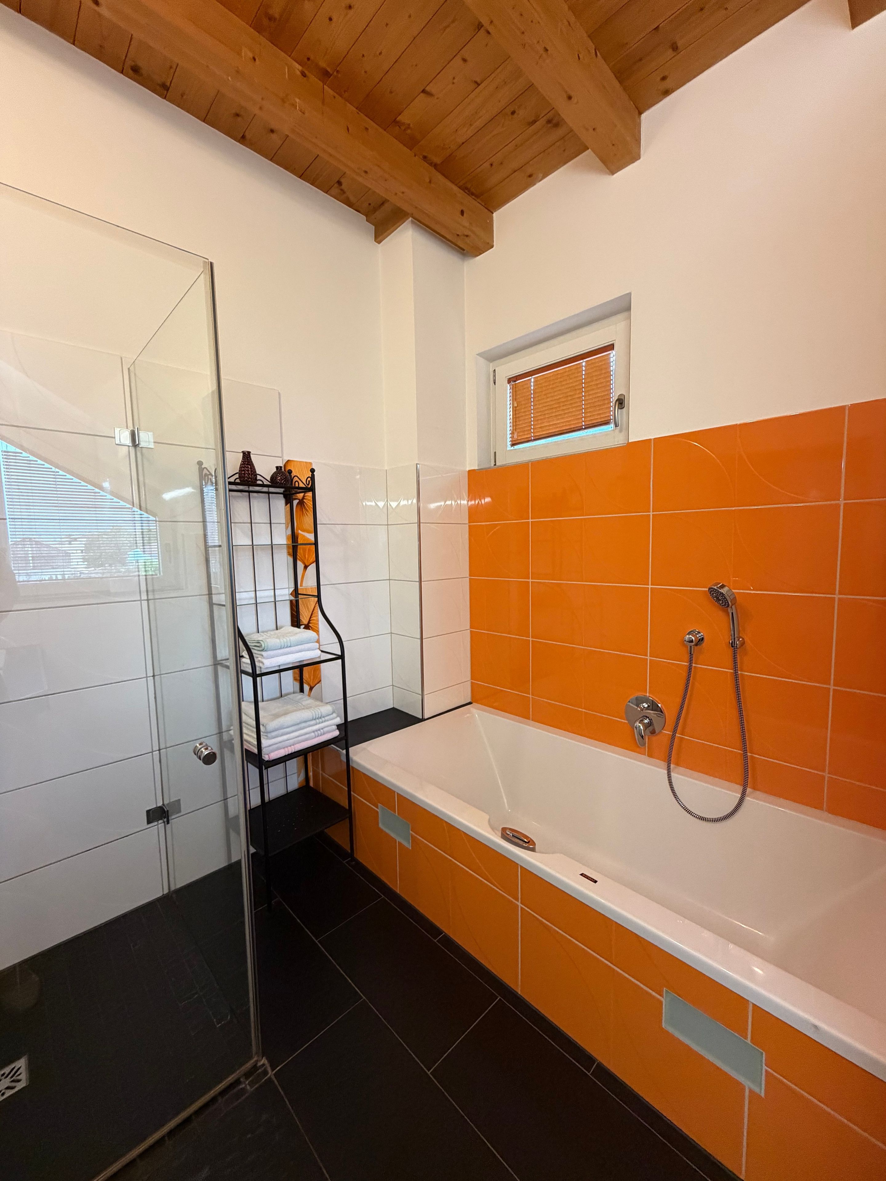 A bathroom with a glass shower enclosure, a white bathtub with an orange tiled wall, and a window with blinds.