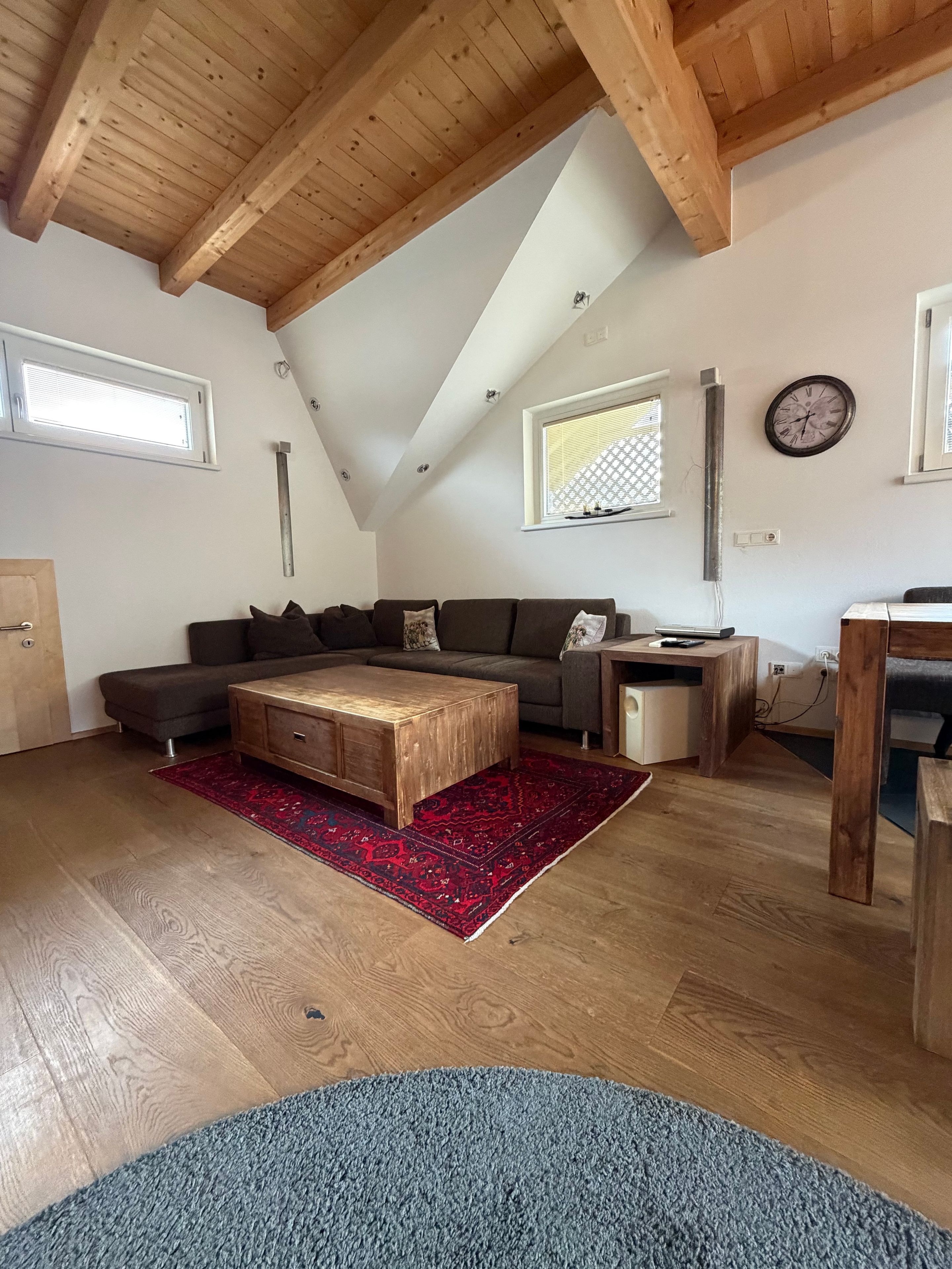 Ein Wohnzimmer im Loft-Stil mit schrägem Dach, Holzbalken, einer Sofagarnitur, einem Couchtisch und einem Teppich.
