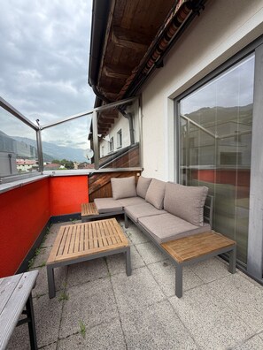 A balcony with a wooden table and a sofa, a view of a building with a red wall, and a large window with blinds.