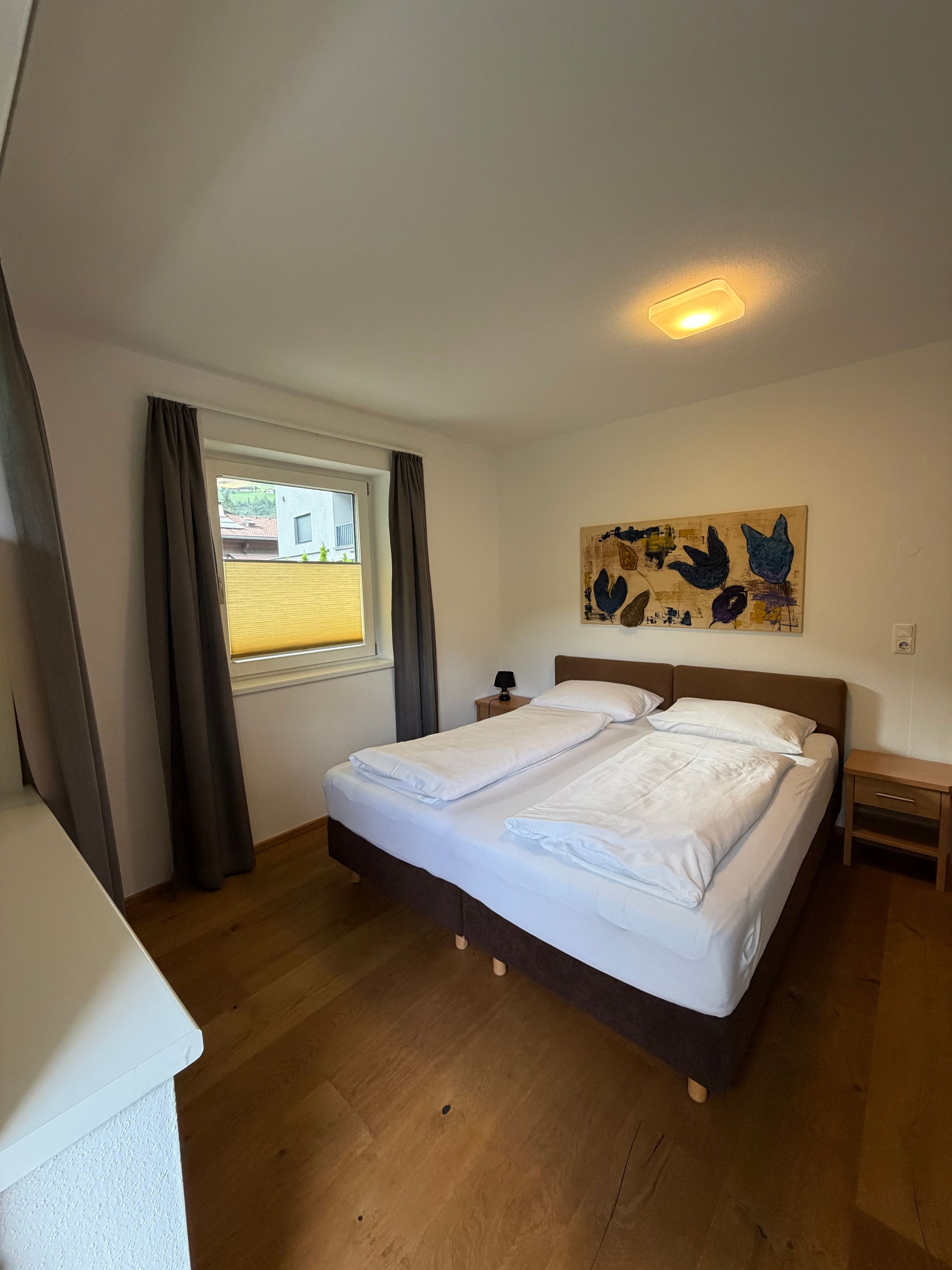 Ein Schlafzimmer mit Bett, einem Fenster mit Vorhängen, einem Nachttisch, einer Lampe und einem Wandbild.