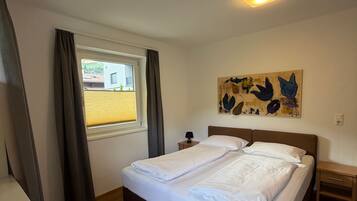 Ein Schlafzimmer mit Bett, einem Fenster mit Vorhängen, einem Nachttisch, einer Lampe und einem Wandbild.