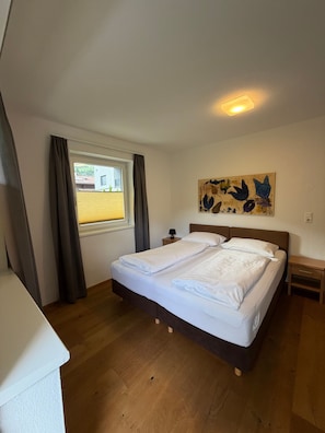 Een slaapkamer met een bed, een raam met gordijnen, een nachtkastje, een lamp en een kunstwerk aan de muur.