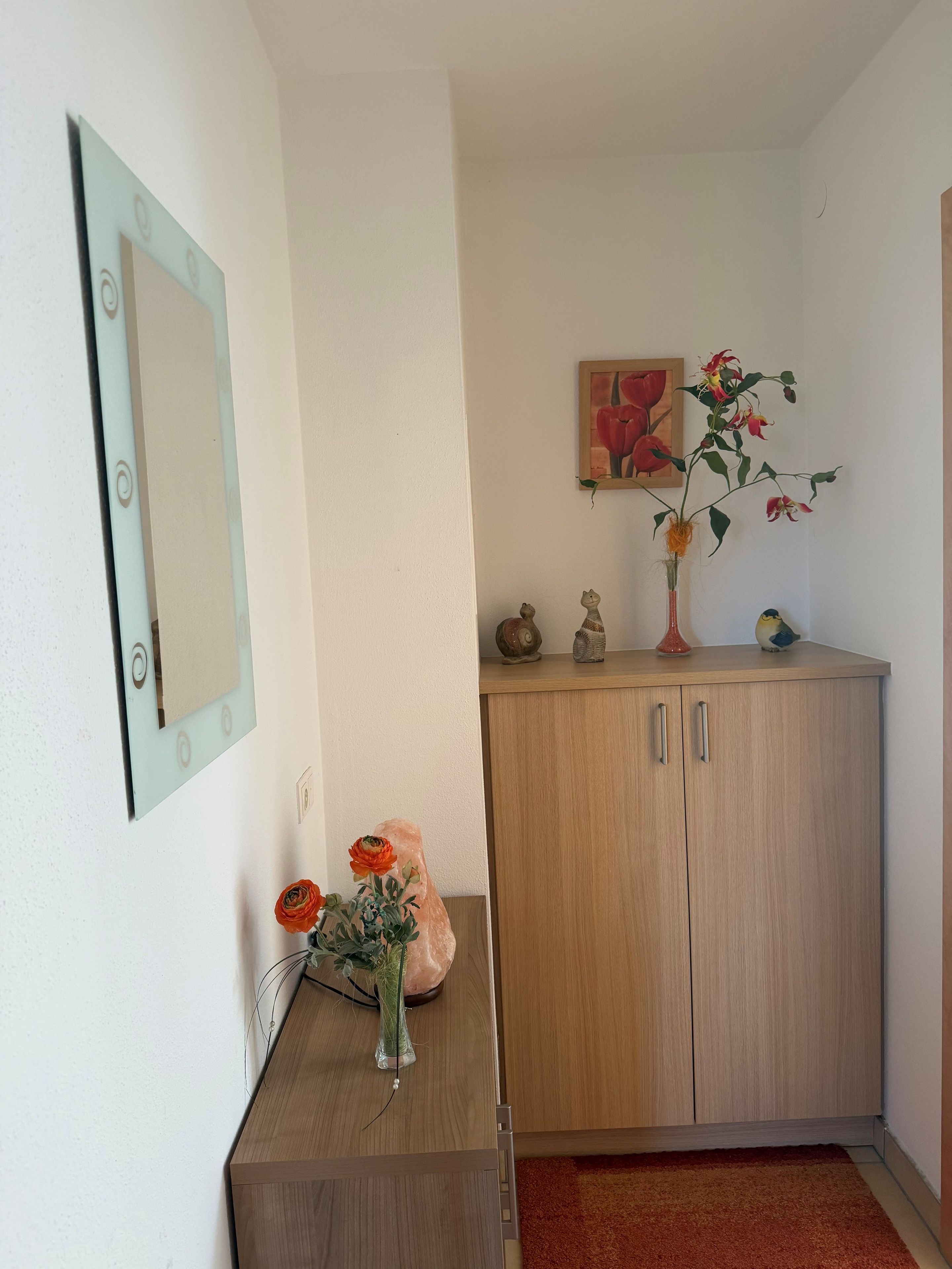 A room with a wooden cabinet, a mirror, a vase with flowers, and a painting on the wall.