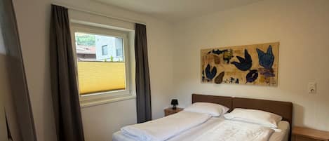 Un dormitorio con cama, ventana con cortinas, mesita de noche con lámpara y una obra de arte en la pared.