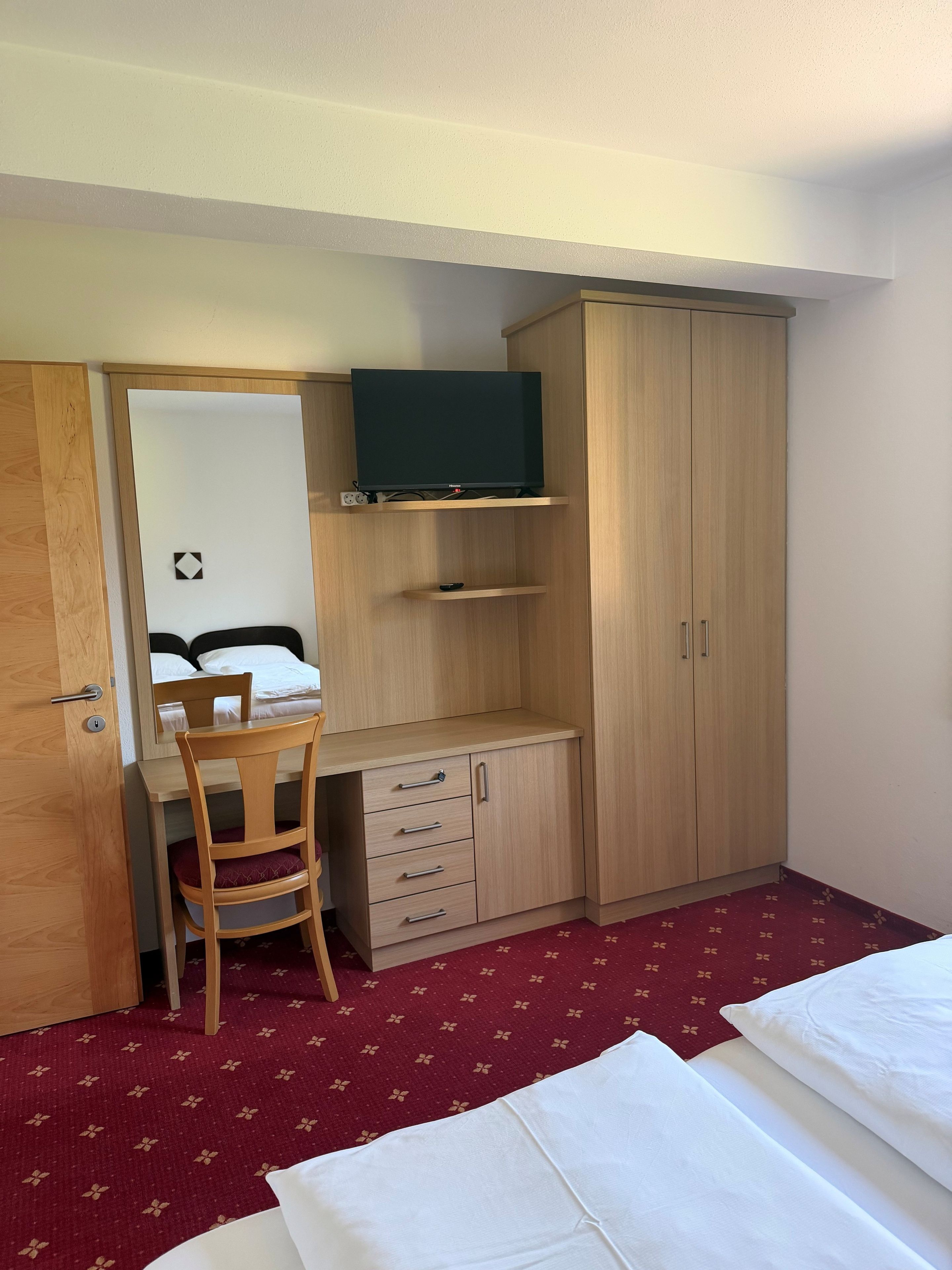 Quarto de hotel pequeno com uma cama, uma escrivaninha com cadeira, um guarda-roupa e uma televisão numa prateleira.