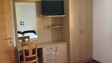 Quarto de hotel pequeno com uma cama, uma escrivaninha com cadeira, um guarda-roupa e uma televisão numa prateleira.