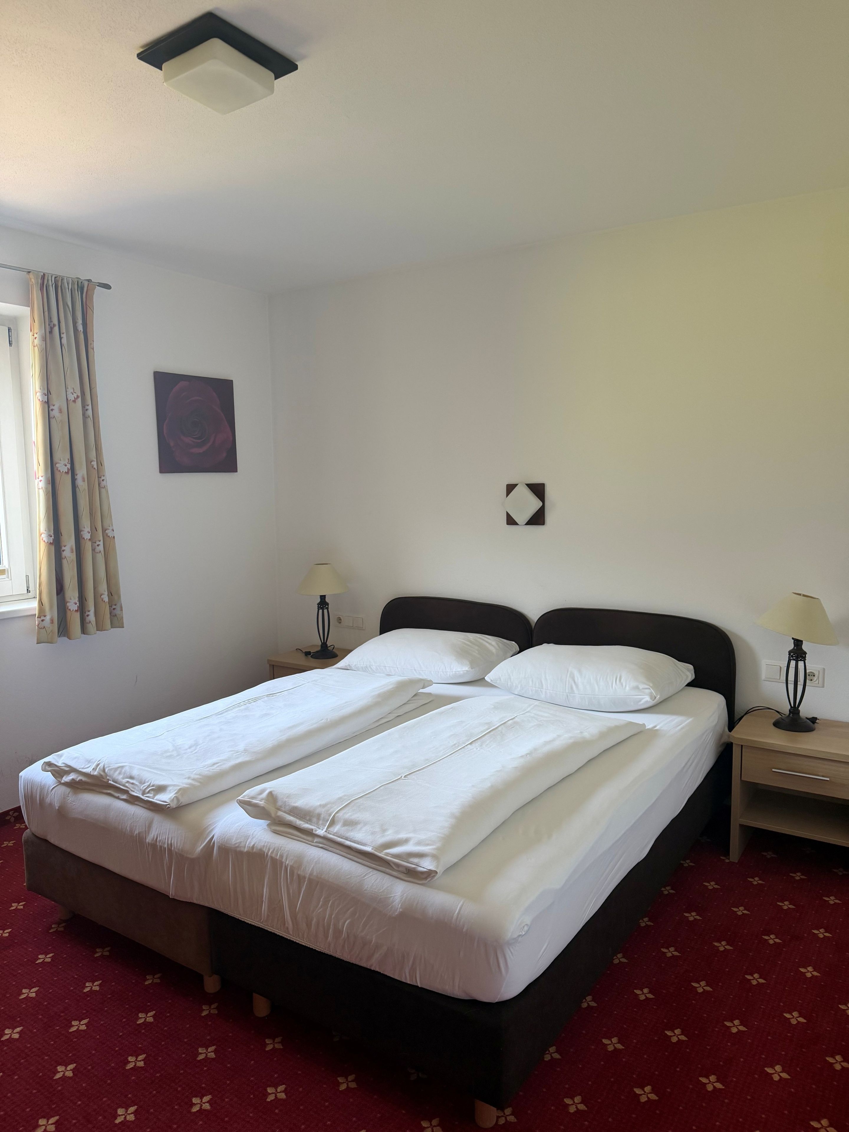 Hotellihuone, jossa on parivuode, kaksi yöpöytää, ikkuna verhoilla, seinälle ripustettu taulu ja kattovalaisin.