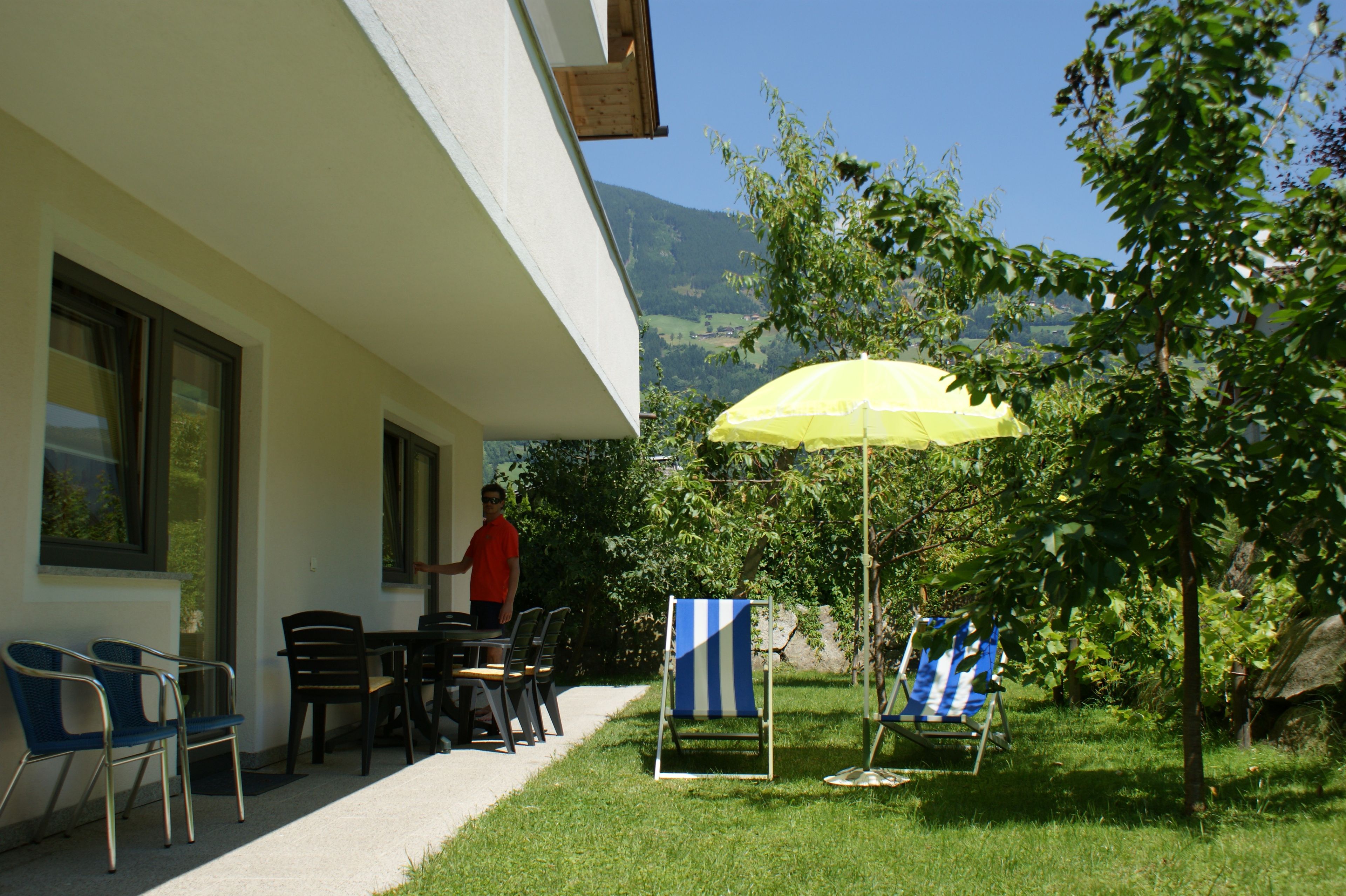 Ein Außenbereich mit Sitzgelegenheit, einem Tisch, Stühlen und einem gelben Sonnenschirm, mit Blick auf einen Berg.
