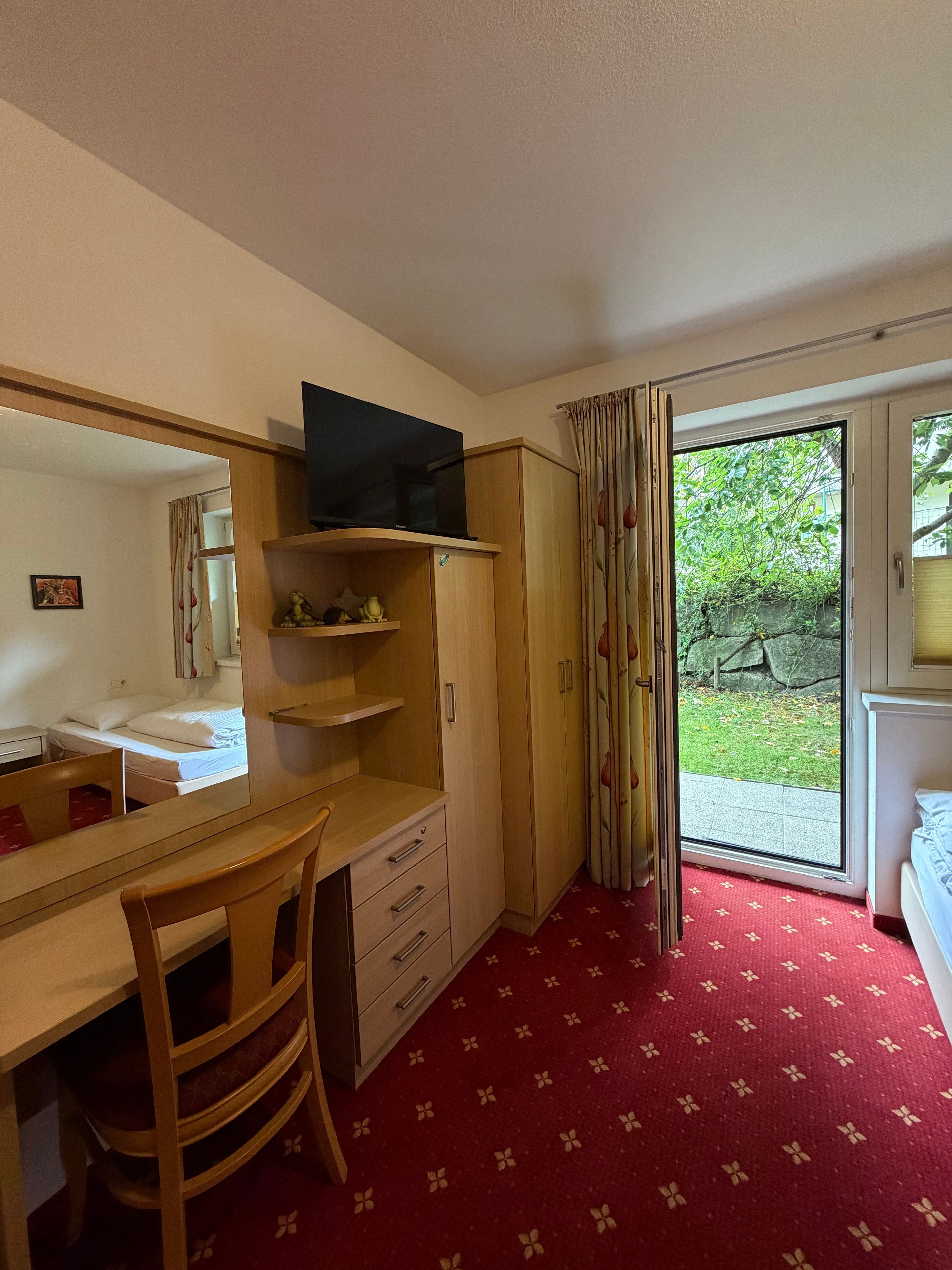 A compact room with a desk, chair, and a television on a stand. There is a wardrobe and a bed with a red patterned carpet.