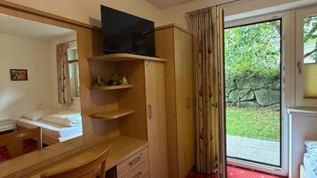 A compact room with a desk, chair, and a television on a stand. There is a wardrobe and a bed with a red patterned carpet.