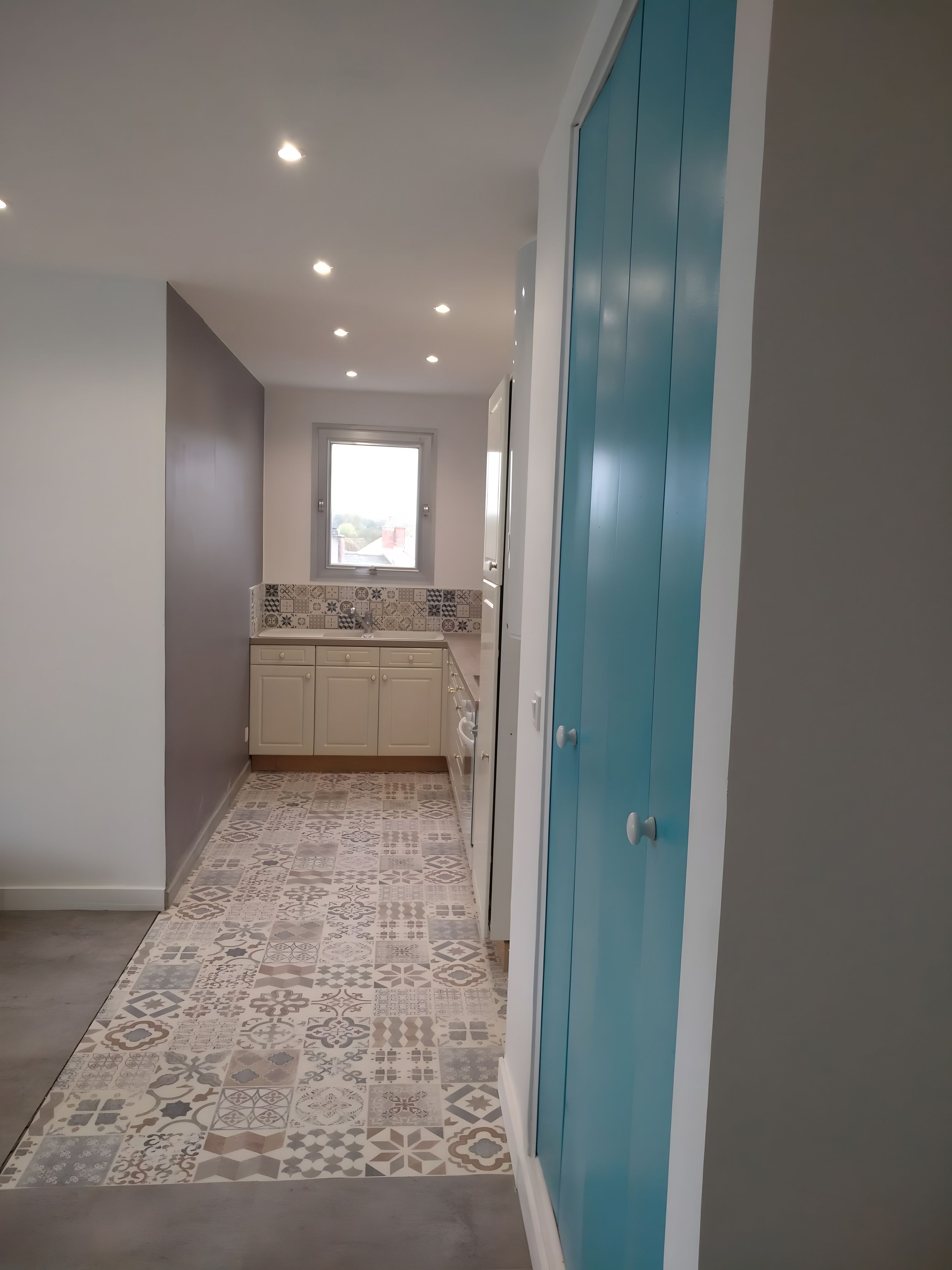 Un couloir étroit avec des carreaux de sol à motifs, une fenêtre donnant sur un paysage agréable et une porte bleue.