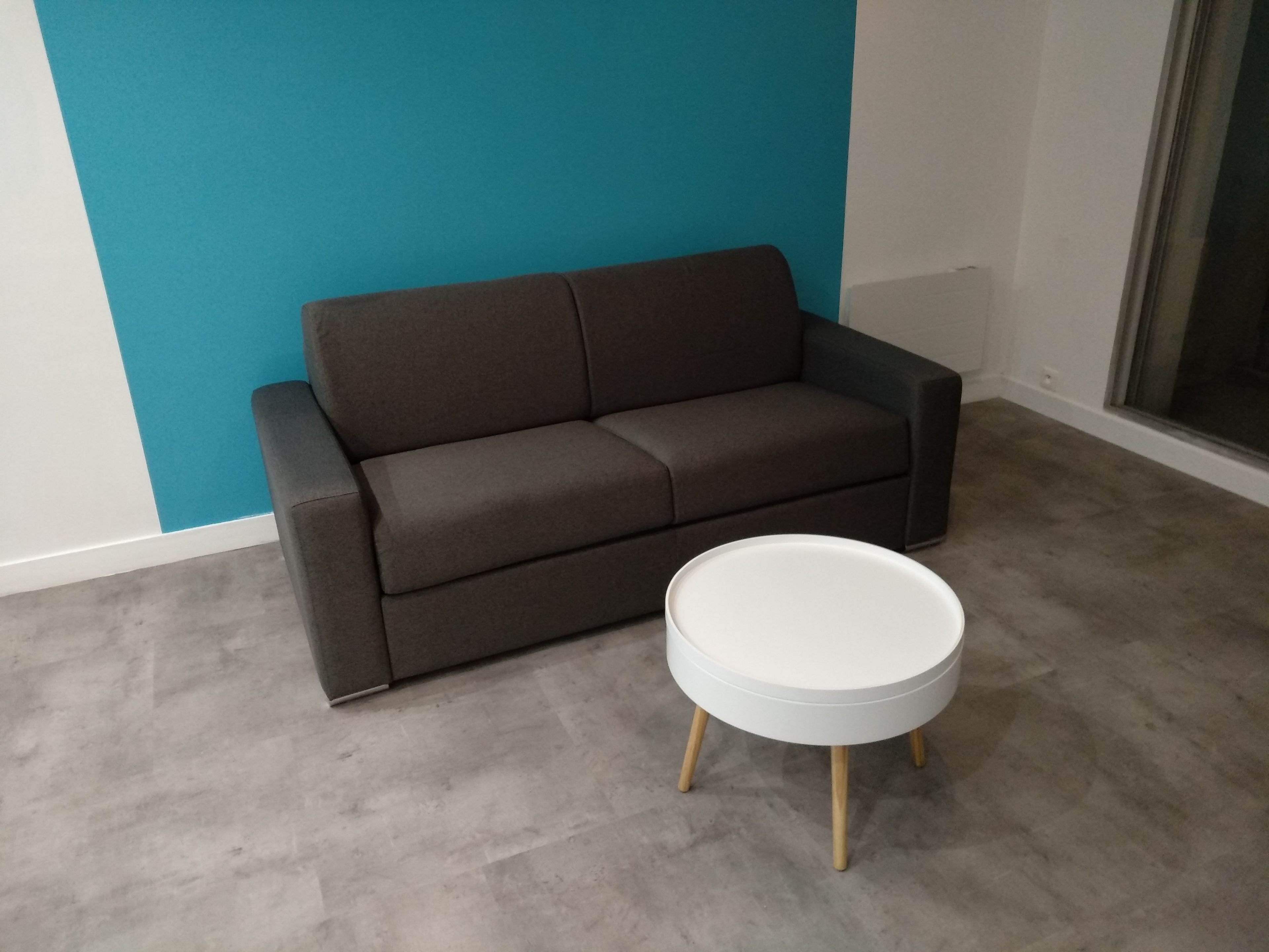 Une pièce avec un canapé gris et une table ronde blanche aux pieds en bois.