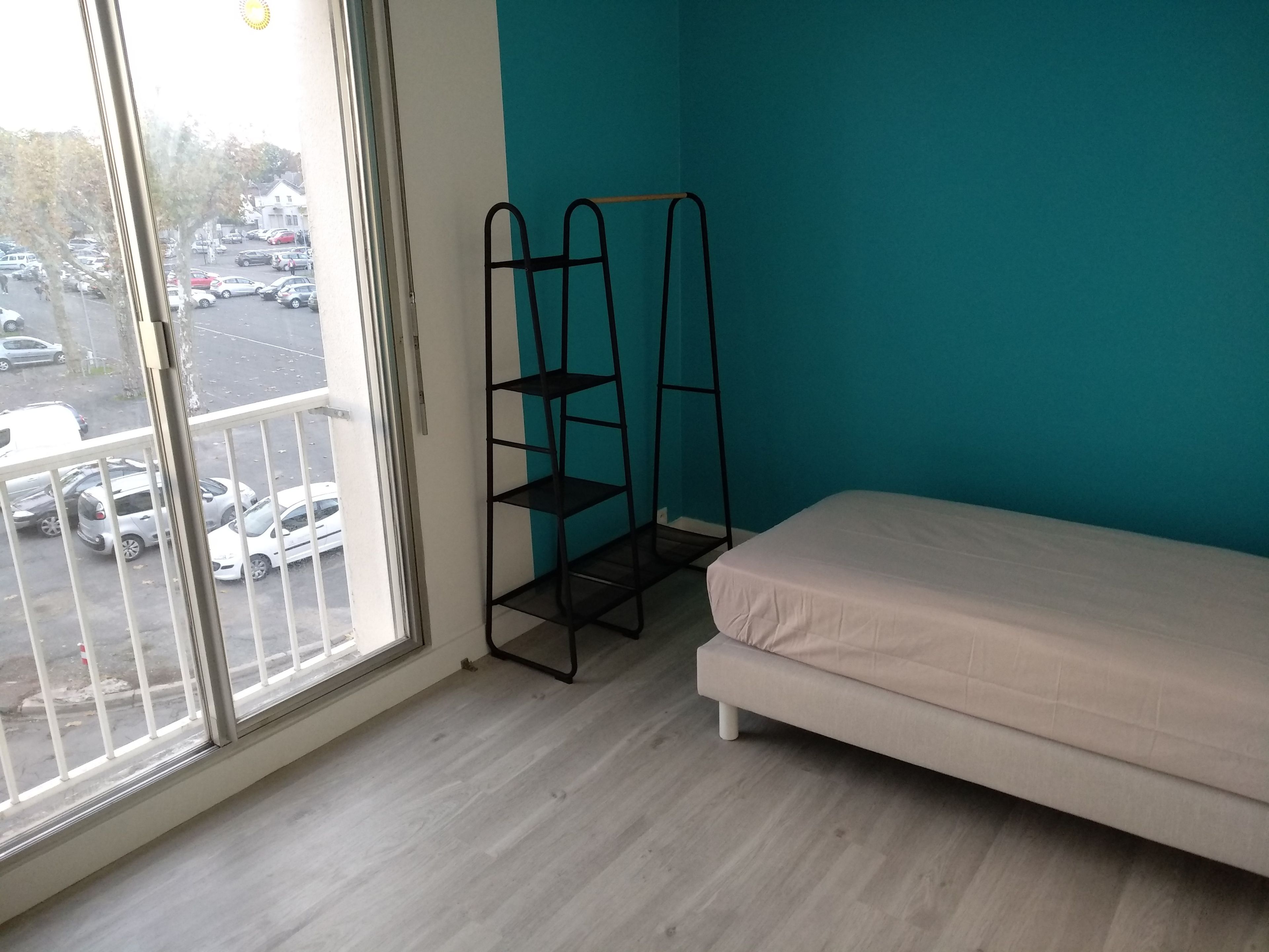 A room with a bed, a window with a view of parked cars, and a metal shelf unit.
