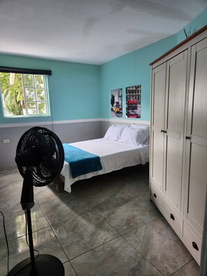Une chambre avec un lit, un ventilateur, une armoire et une fenêtre donnant sur des arbres.