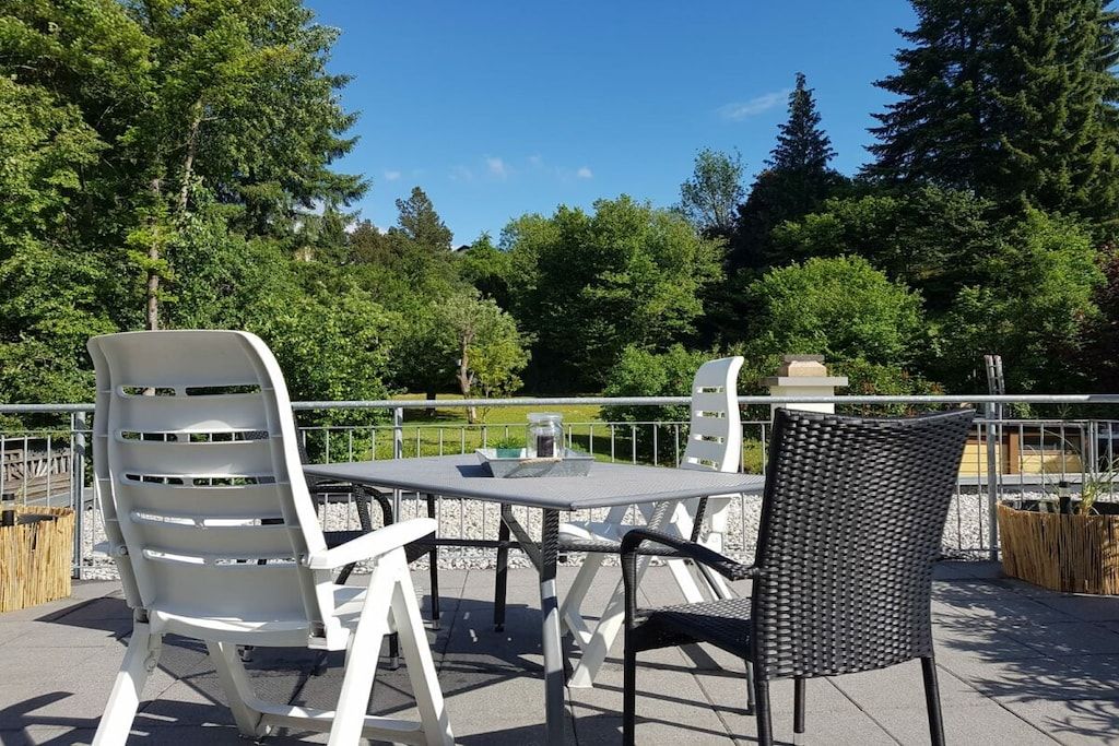 An outdoor patio with a table and chairs, surrounded by a metal railing, with trees in the background.