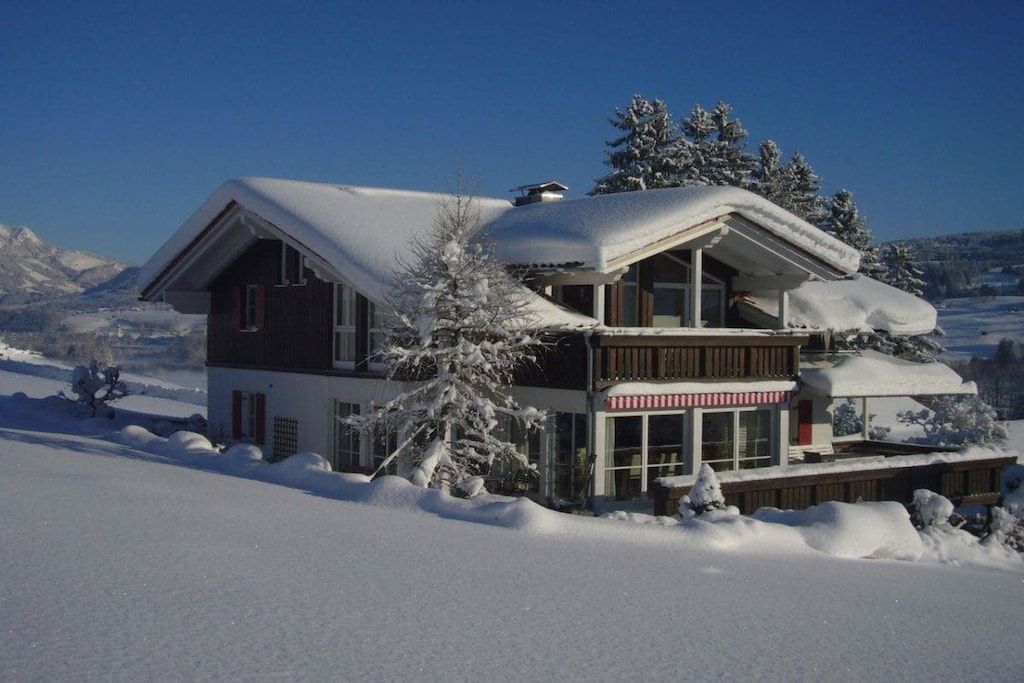 Een chalet met twee verdiepingen, een met sneeuw bedekte kap en een balkon, omgeven door een besneeerd landschap en bergen.