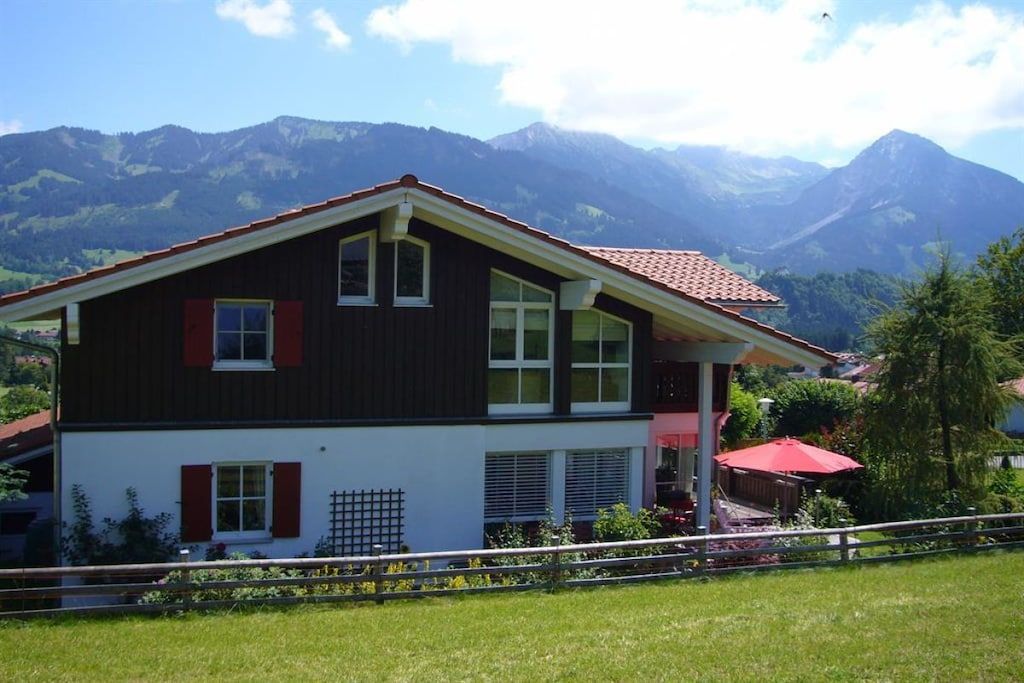 A two-story house with a balcony, surrounded by a fence, and mountains in the background.