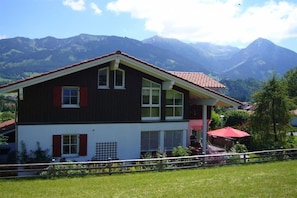 A two-story house with a balcony, surrounded by a fence, and mountains in the background. - Holiday apartment with 1 bedroom (Fischen)