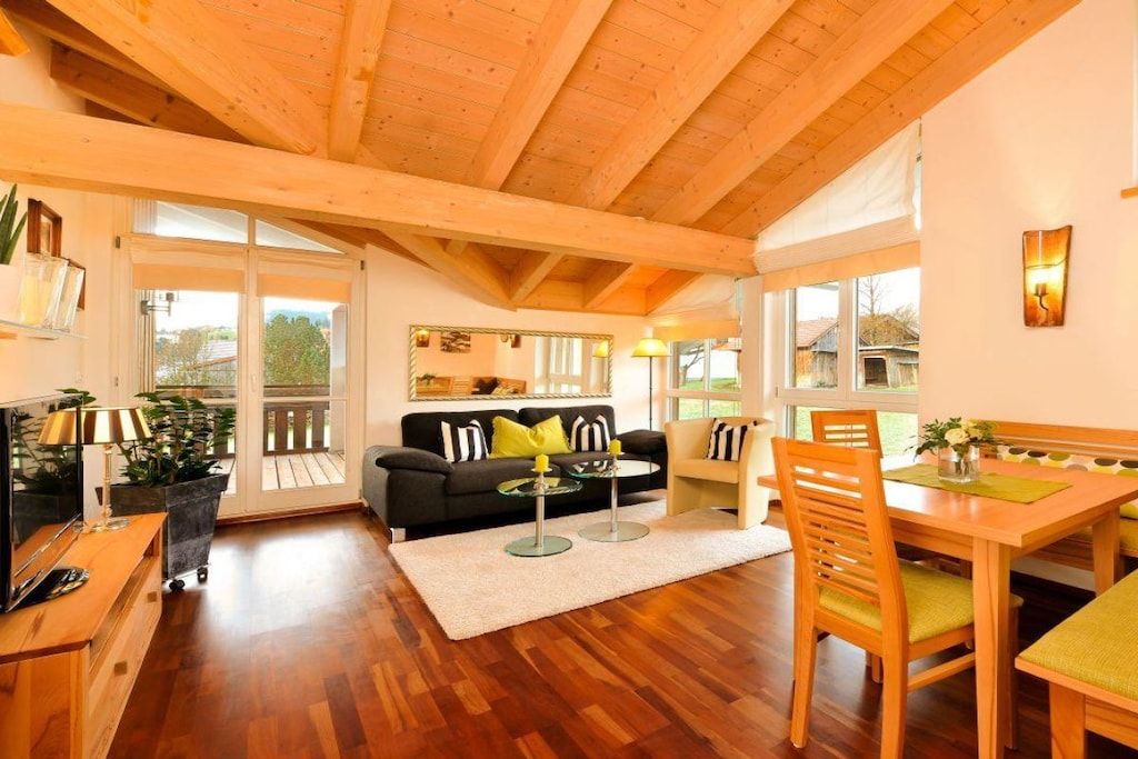 A spacious living area with a wooden ceiling, a sofa, a dining table with chairs, and a large window providing a view of the outdoors.