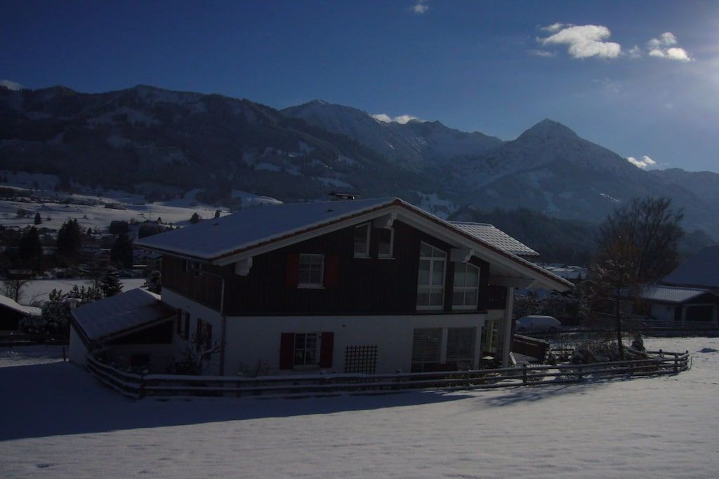 A two-story house with a sloped roof, surrounded by a snow-covered landscape and mountains in the background.