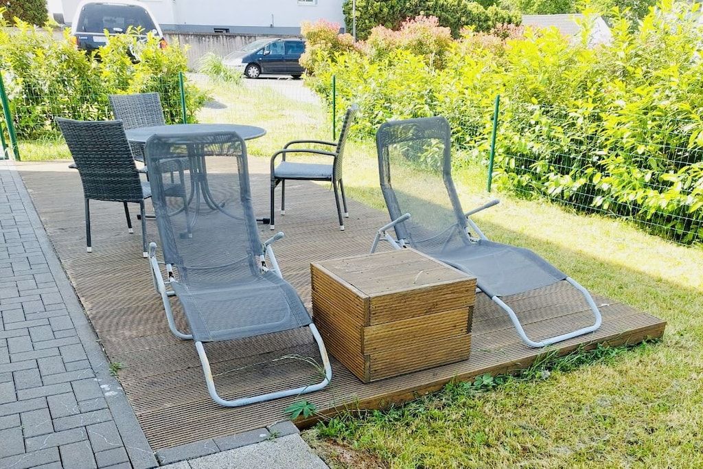 Un área de estar al aire libre con sillas de metal y una mesa, rodeada por una terraza de madera y una zona con césped.
