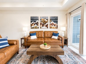 A living room with a brown leather sofa, two side tables with lamps, a coffee table with a plant, and a patterned rug. - New Pigeon Forge Condo | Mt. Views (Pigeon Forge)