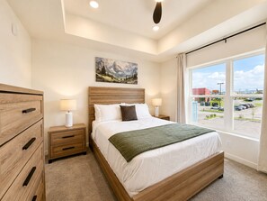 A bedroom with a large bed, wooden headboard, bedside tables with lamps, a painting on the wall, and a view of a parking lot through a large window. - New Pigeon Forge Condo | Mt. Views (Pigeon Forge)