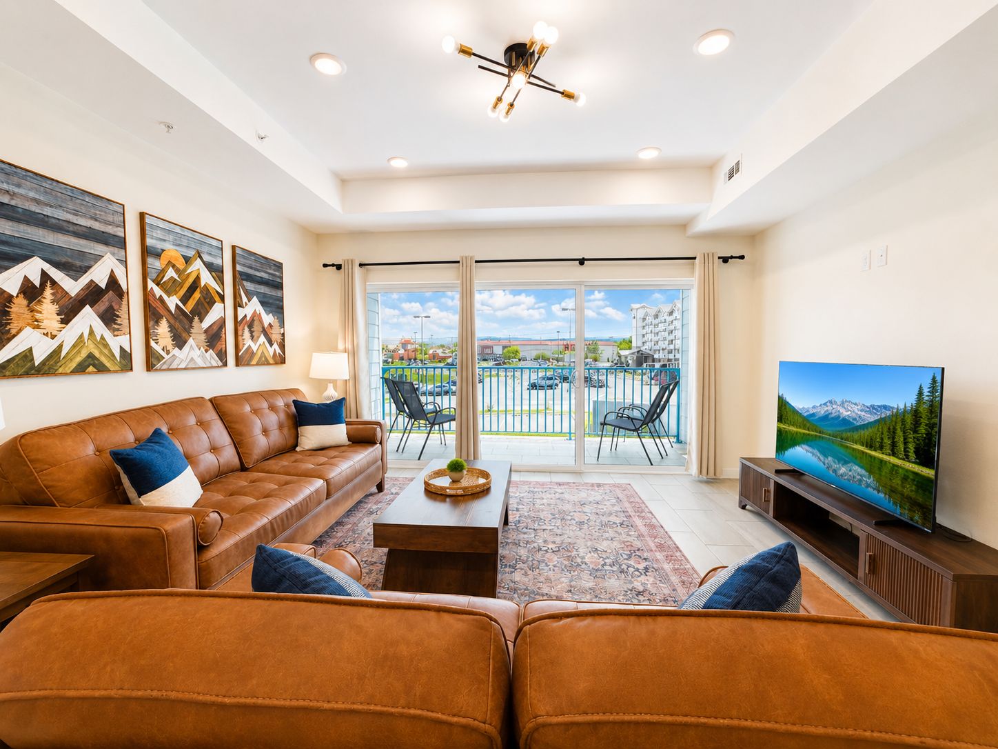 A living room with a brown leather sofa, a coffee table, and a flat-screen TV. There are three framed artworks on the wall and a balcony with a view of the outside.