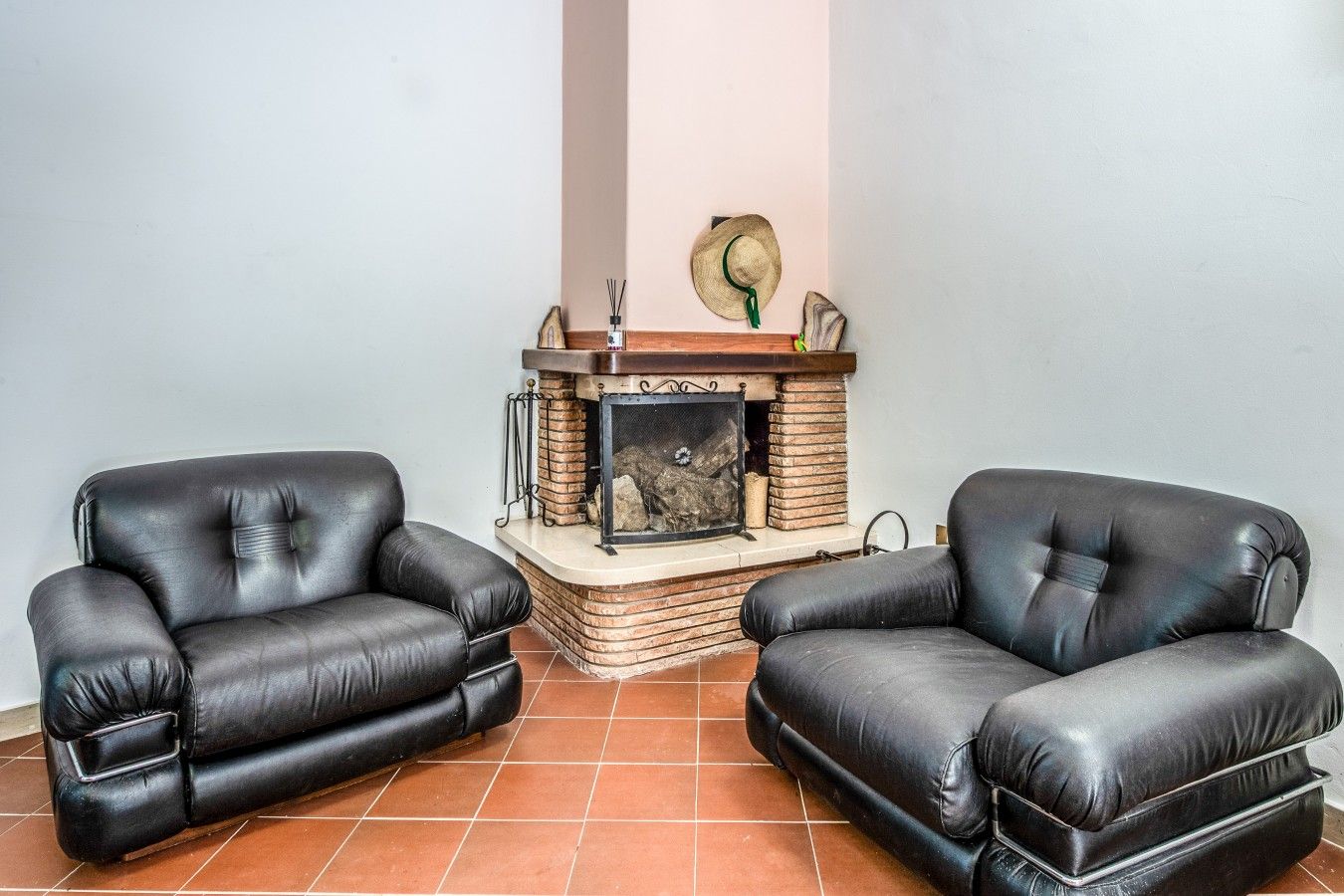 Une pièce avec deux fauteuils en cuir noir, une cheminée avec un manteau en pierre et un sol carrelé.