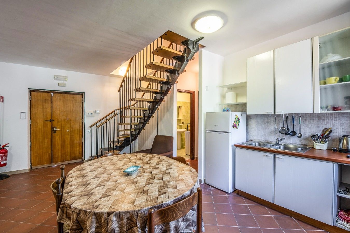 A kitchen with a built-in stove, a sink, and cabinets. There is a wooden dining table with chairs. A spiral staircase leads to an upper level.