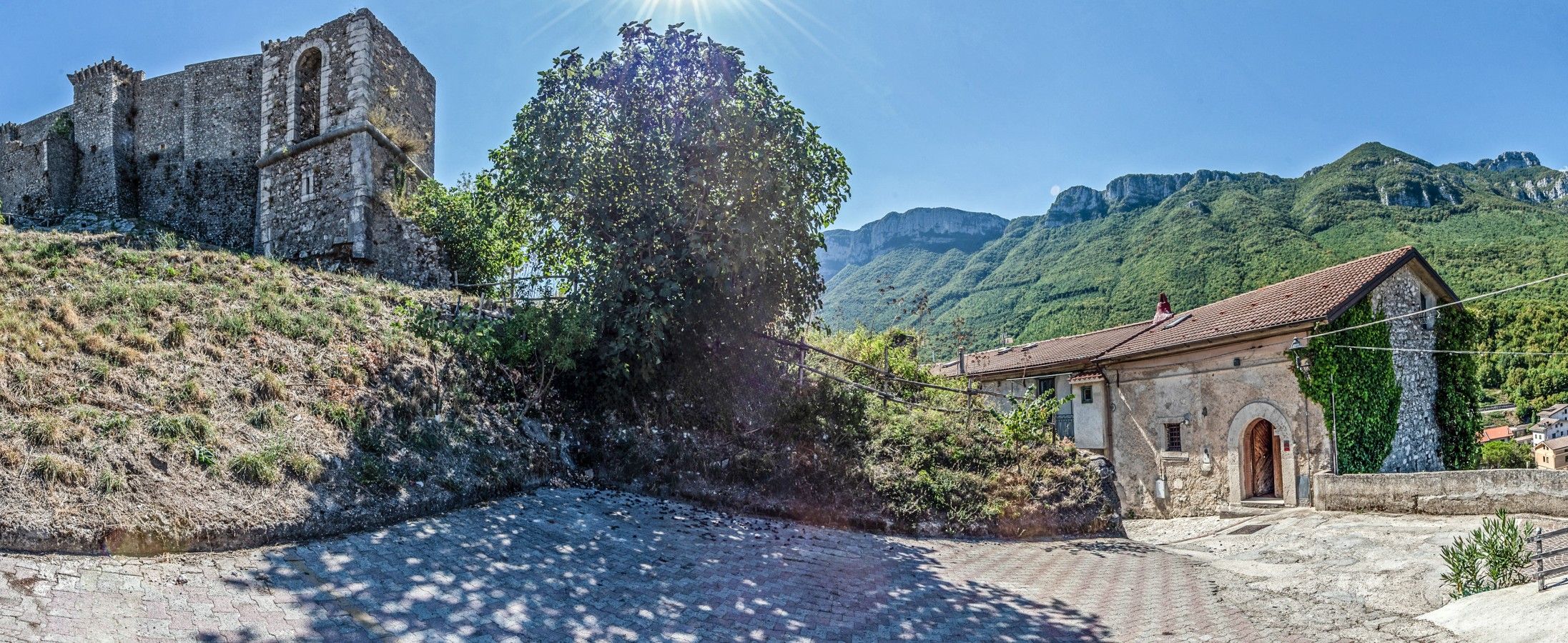A stone building with a tiled roof, a large tree in front, and a mountainous backdrop.