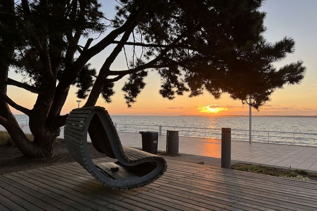 Eine Holzterrasse mit einer gebogenen Bank, einem Baum und Blick auf den Sonnenuntergang über dem Wasser.