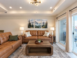 A living room with a brown leather sofa, a wooden coffee table, and two wall-mounted lamps. There are three framed paintings above the sofa depicting mountain landscapes. - New Pigeon Forge Condo | Island View (Pigeon Forge)