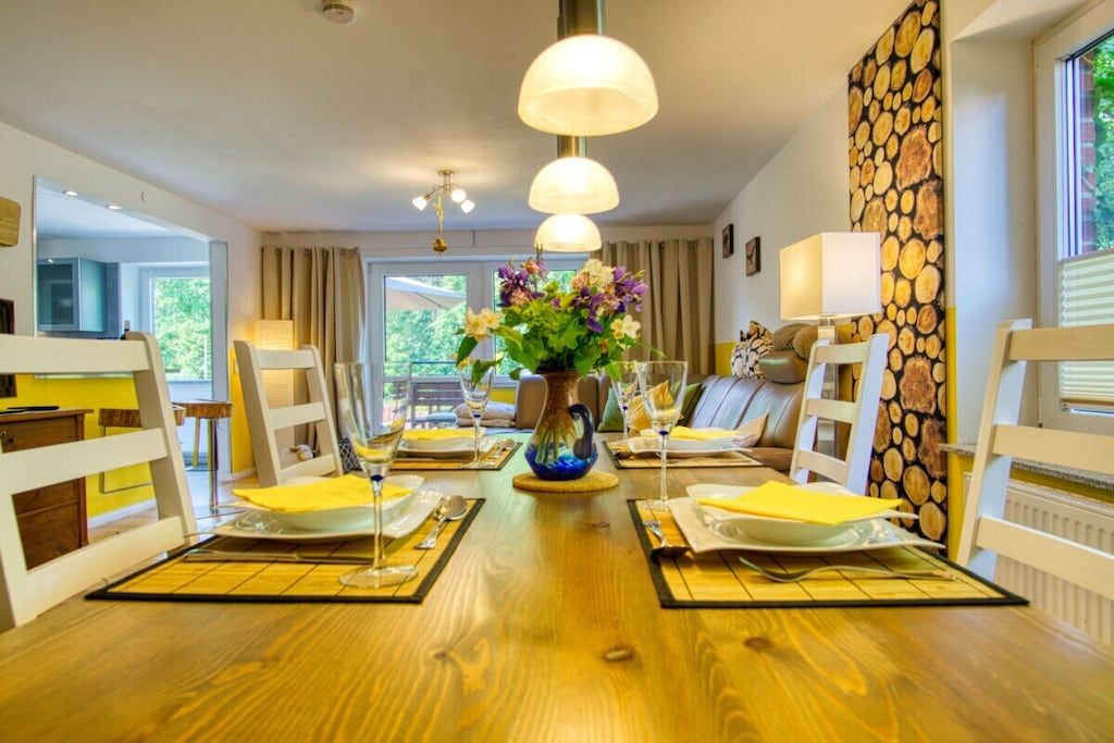 A dining area with a wooden table set for four, surrounded by white chairs, with a vase of flowers in the center.