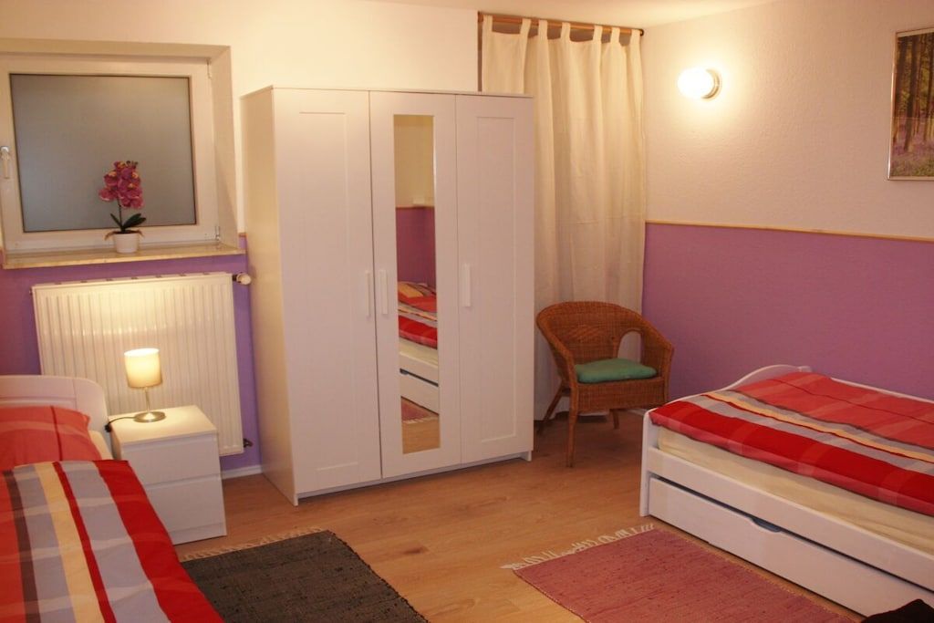 Una camera da letto con un letto, un armadio, una sedia, un comodino, una lampada, una finestra con tende e un termosifone.