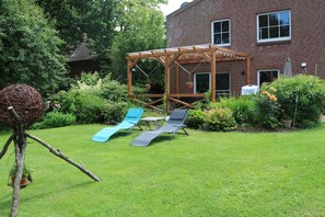 Een achtertuin met een houten pergola, twee lounge stoelen, een schommel en een bakstenen gebouw op de achtergrond.