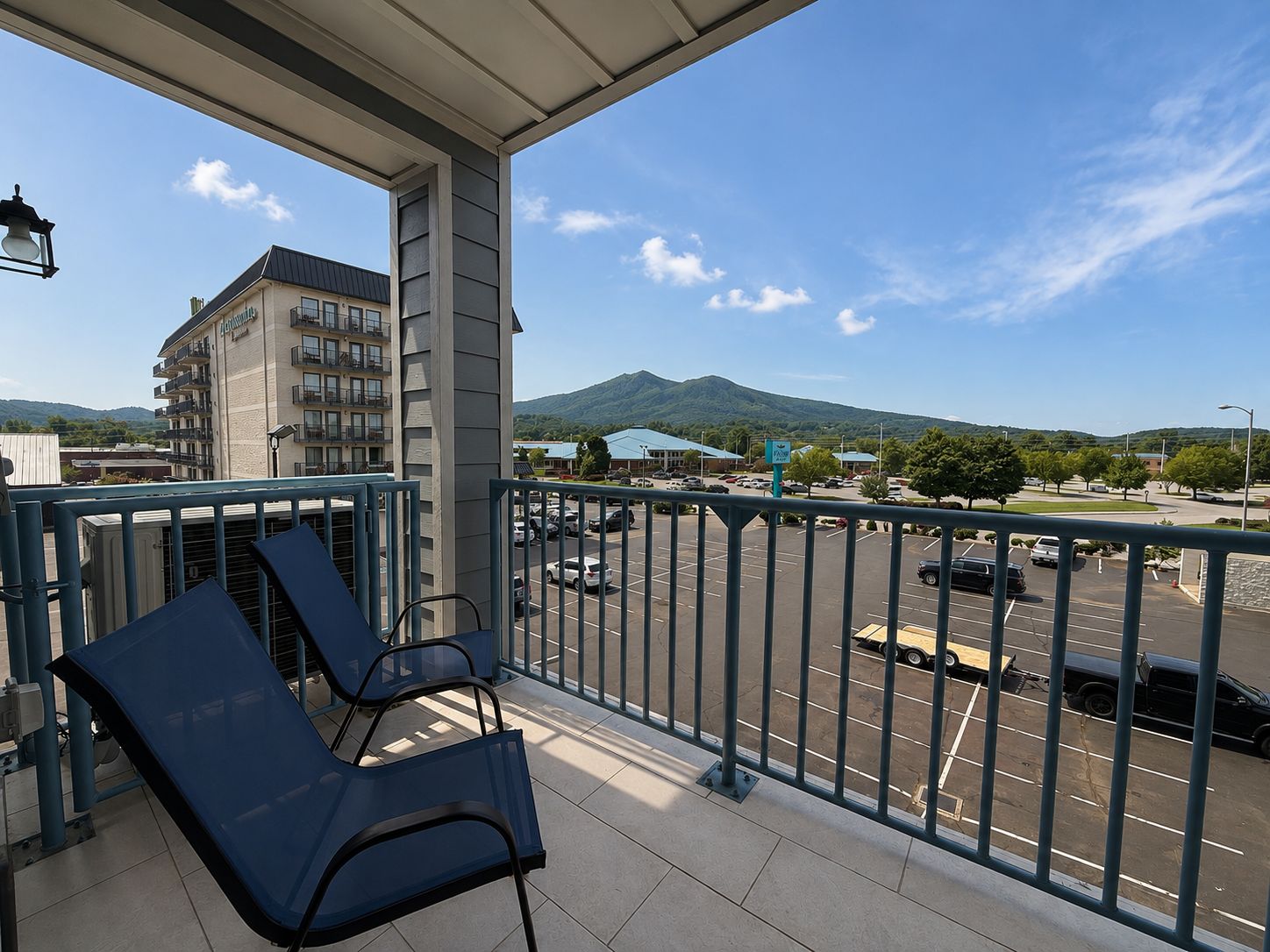 En balkon med to stole, udsigt til en parkeringsplads, en bygning med balkoner og et bjerg i baggrunden.