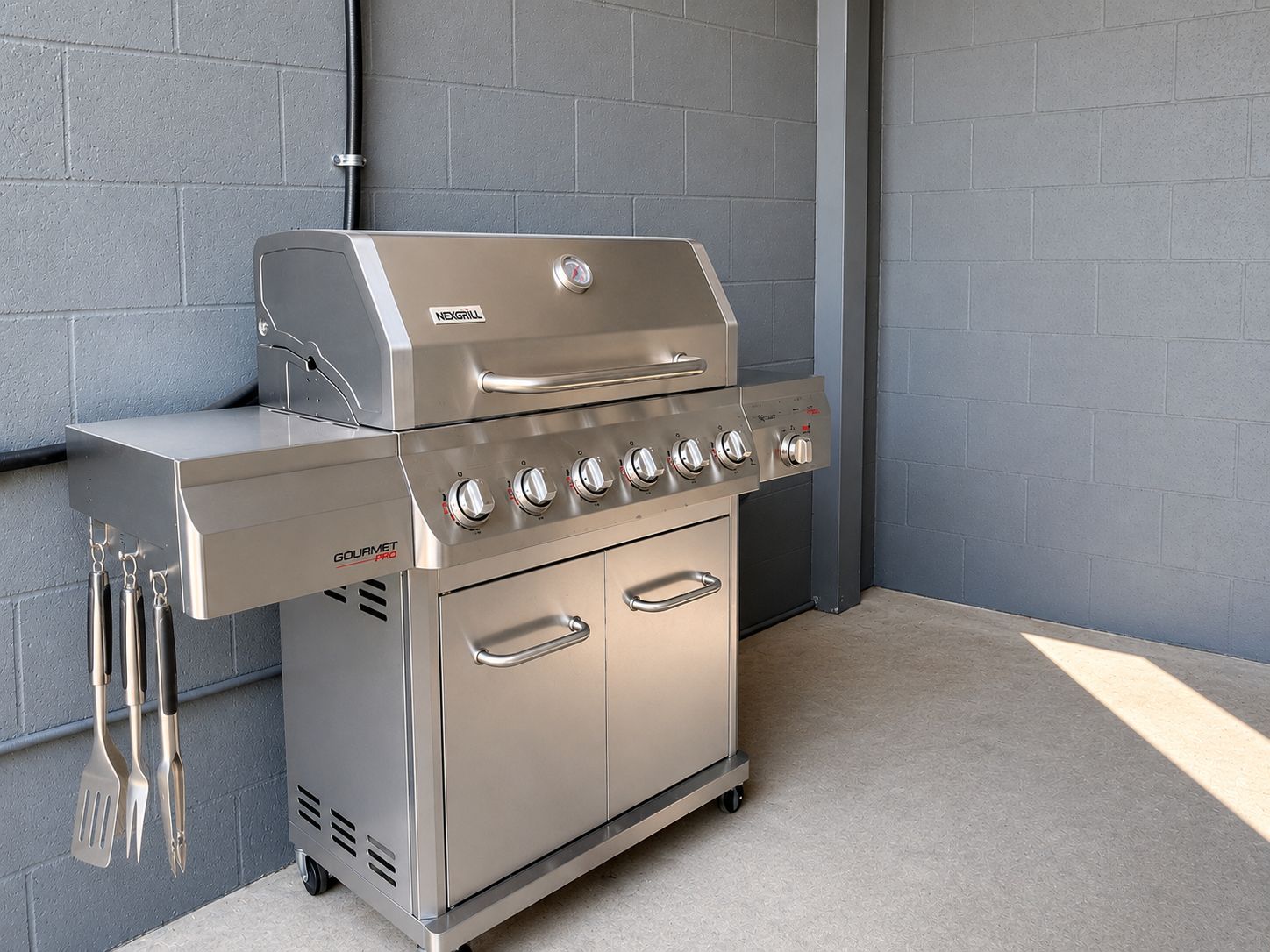 A stainless steel outdoor grill with multiple burners and a side cabinet, set against a grey brick wall.