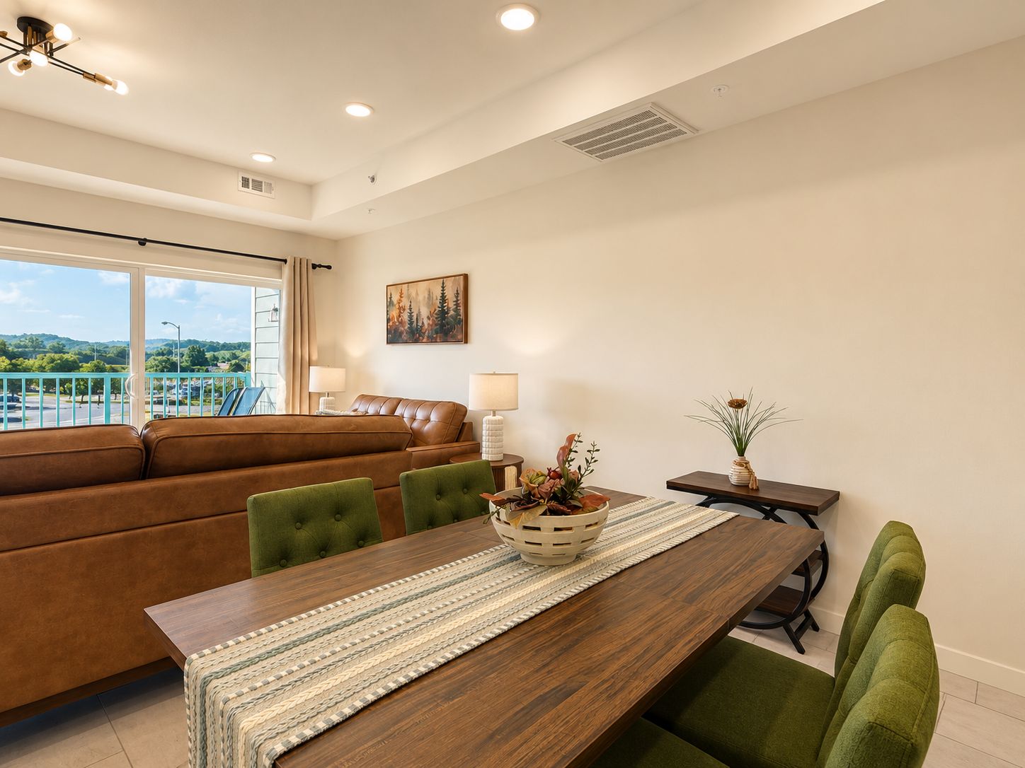 Una sala de estar moderna con una mesa de comedor de madera, sillas tapizadas en verde, un sofá de cuero marrón y vistas a un paisaje verde a través de grandes ventanales.