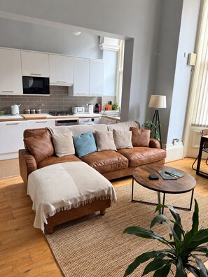 A modern living room with a brown leather sofa, a coffee table, and a kitchen in the background. - Historic Hull Old Town Apartment (Kingston upon Hull)