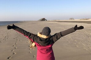 Eine Person steht auf einem sandigen Strand, die Arme geöffnet, und blickt auf ein Gebäude in der Ferne.