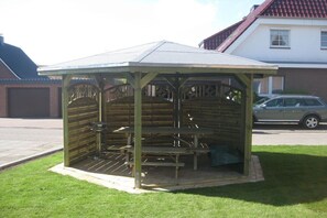 Ein Pavillon mit Dach und Bänken, umgeben von Gras, mit einem Gebäude und einem Auto im Hintergrund.