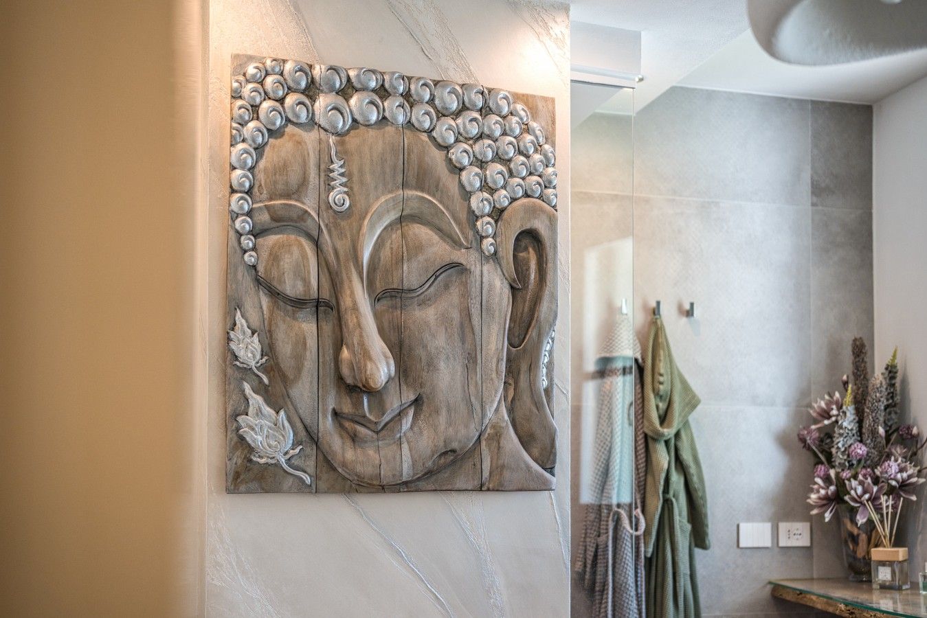 A wall-mounted artwork of a Buddha's face with a serene expression, a towel rack with folded towels, and a vase with dried flowers.
