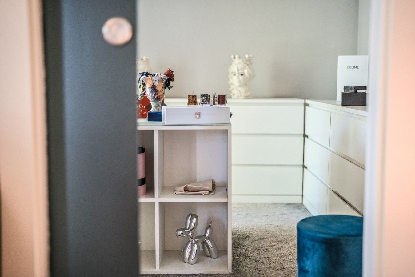 Une pièce avec une commode blanche, une étagère ornée d’objets décoratifs et un pouf bleu.